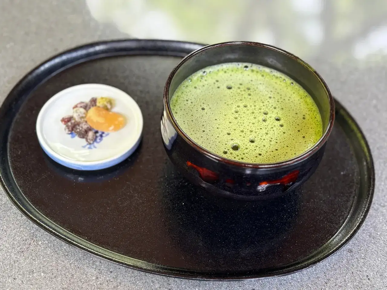 Zielona, spieniona matcha w czarnej miseczce, obok talerzyk z bakaliami. Czy matcha pobudza? Ten napój z pewnością doda energii!