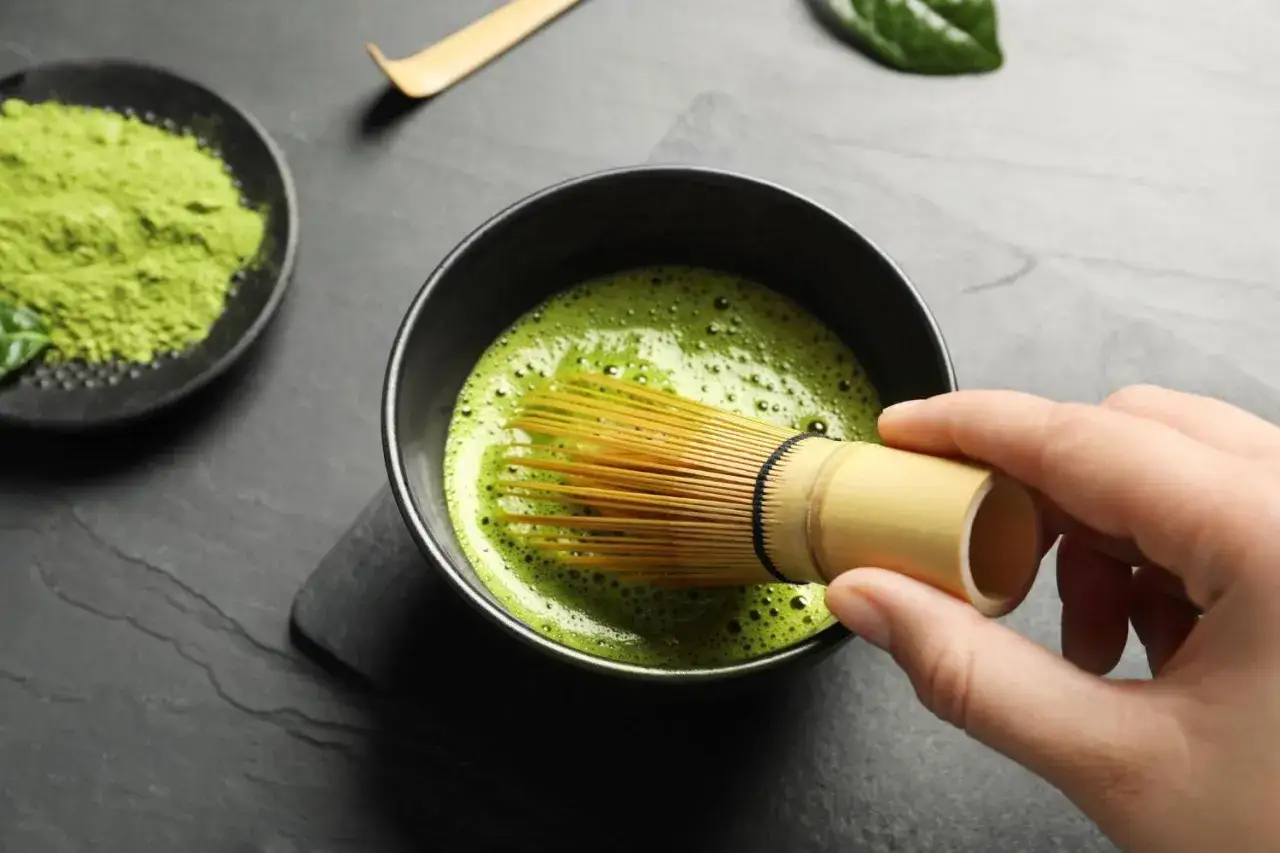 Przygotowanie matcha na co dzień. Zielony proszek, bambusowa miotełka i spieniony nap&oacute;j w czarnej miseczce.