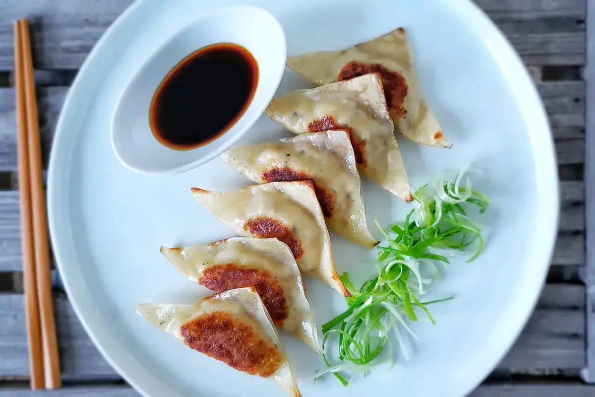 Siedem złocistych pierożków gyoza ułożonych na talerzu, obok sosu sojowego i szczypiorku.