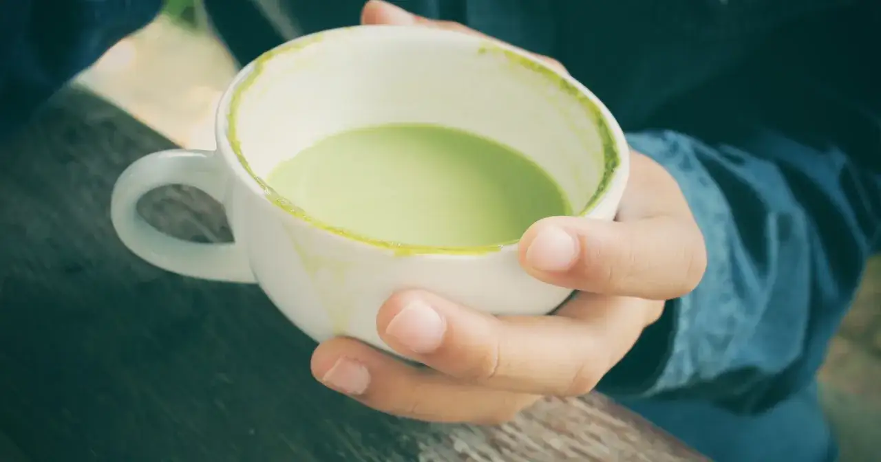 Dłonie trzymają biały kubek z zieloną matchą. Czy matcha pobudza? Ten napój może dodać energii.