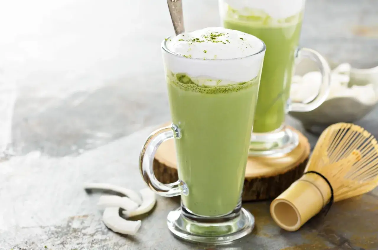 Dwa szklane kubki z zielonym, kremowym napojem i białą pianką. Obok bambusowa miotełka do matchy i wiórki kokosowe. Tak smakuje matcha latte.