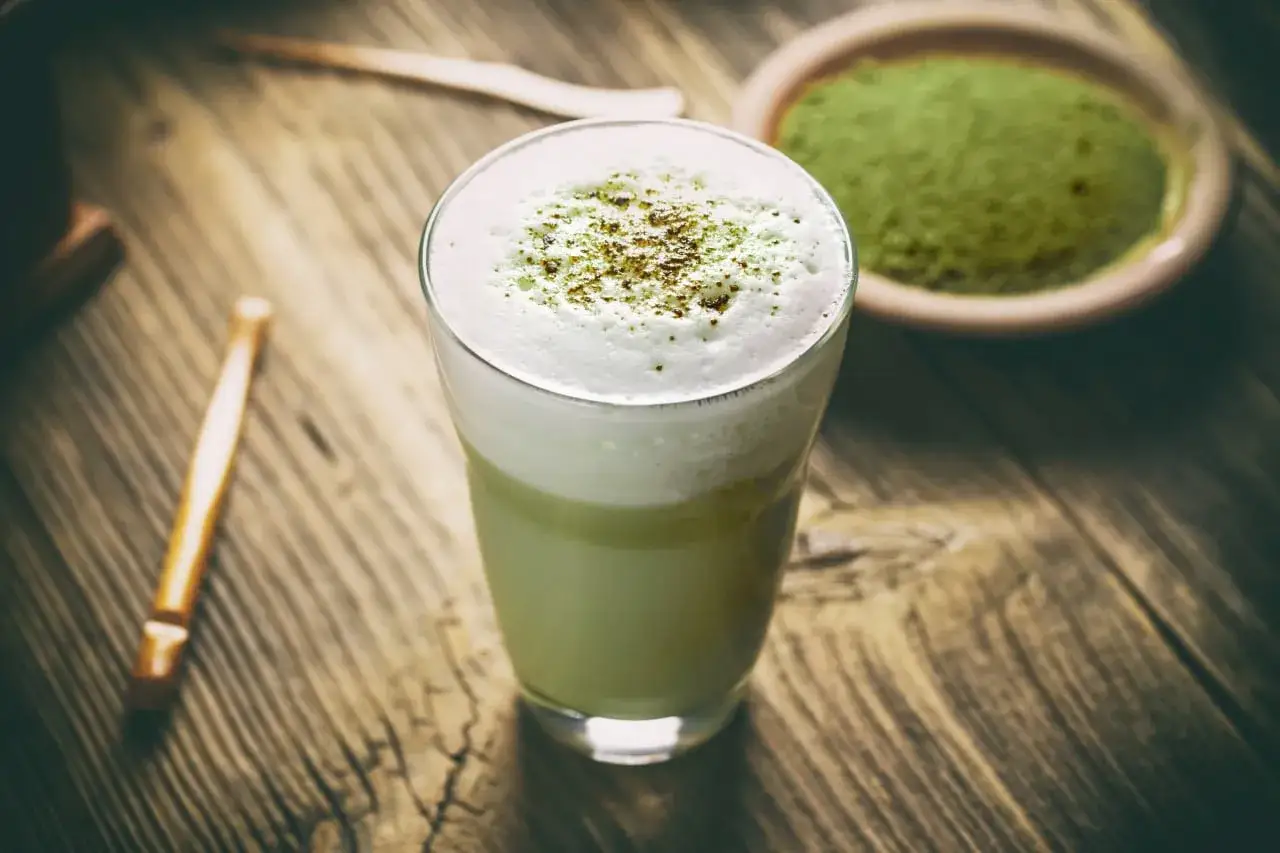 Zielony, spieniony napój w szklance, posypany pyłkiem. Obok miseczka z zielonym proszkiem. To właśnie matcha latte, co to za napój?