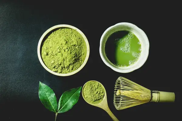 Miska z zielonym proszkiem matcha, filiżanka napoju, drewniana łyżka i bambusowa miotełka.