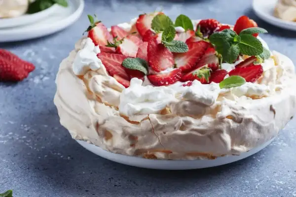 Pyszny tort bezowy z mascarpone i bitą śmietaną, ozdobiony świeżymi truskawkami i miętą.