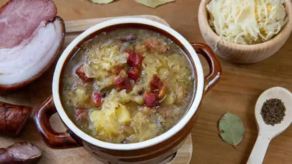 Gęsty, aromatyczny kapuśniak jak u babci, z kawałkami boczku i ziemniaków, podany w glinianym garnuszku. Obok miseczka kiszonej kapusty i przyprawy.