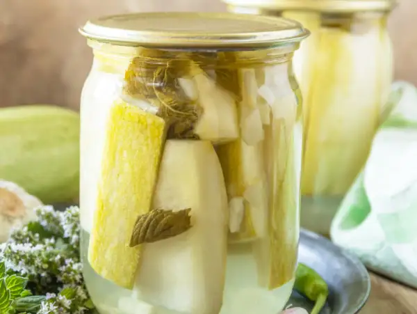 Słoik z cukinią marynowaną po francusku, z dodatkiem ziół i czosnku. Obok leży świeża cukinia.