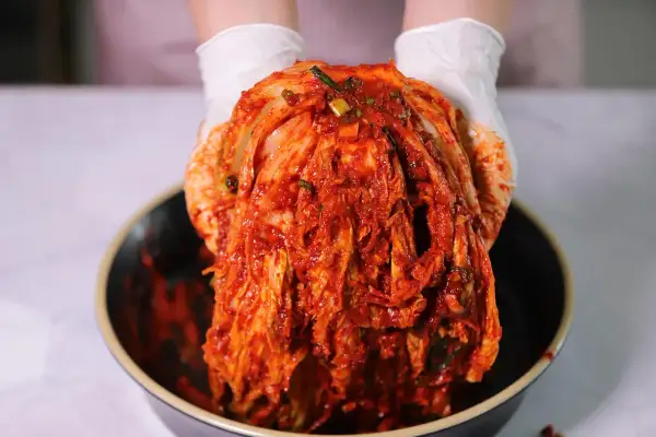 Ręce w białych rękawiczkach trzymają całą, czerwoną kapustę kimchi nad miską. Gdzie kupić kapustę kimchi?