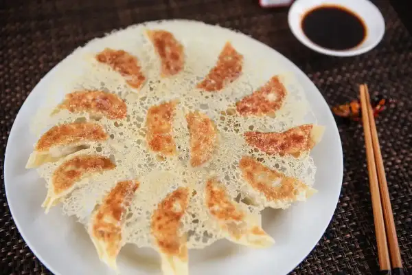 Chrupiące pierożki gyoza ułożone na talerzu, z delikatną, złotą panierką z ciasta. Obok miseczka sosu sojowego i pałeczki.