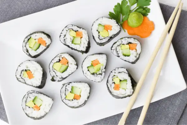 Jak ugotować ryż do sushi? Na talerzu ułożone rolki sushi z awokado, ogórkiem i marchewką. Obok pałeczki i dekoracja z wasabi.