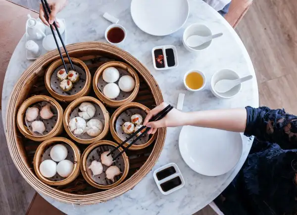 Różnorodne dim sum w bambusowych parownikach, gotowe do degustacji. Ręka z pałeczkami wybiera jeden z przysmaków.